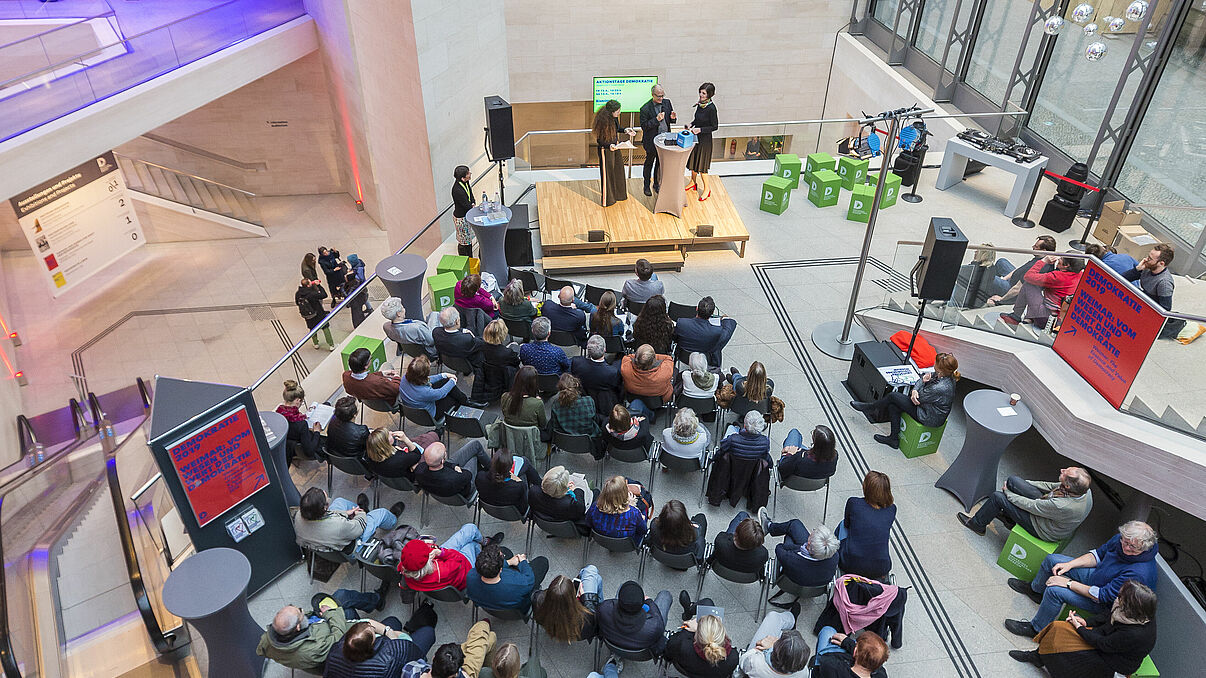 Menschen sitzen vor einer Bühne und hören drei Personen bei einem Gespräch zu.