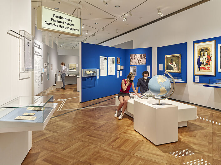 Auf dem Bild sind Personen in der Ausstellung zu sehen. Im Hintergrund sind blaue Wände zu sehen, an denen Objekte hängen. Im Vordergund sitzen Personen und gucken sich Objekte an. Ein Schild mit der Aufschrift "Passkontrolle" ist links im Bild zu sehen.