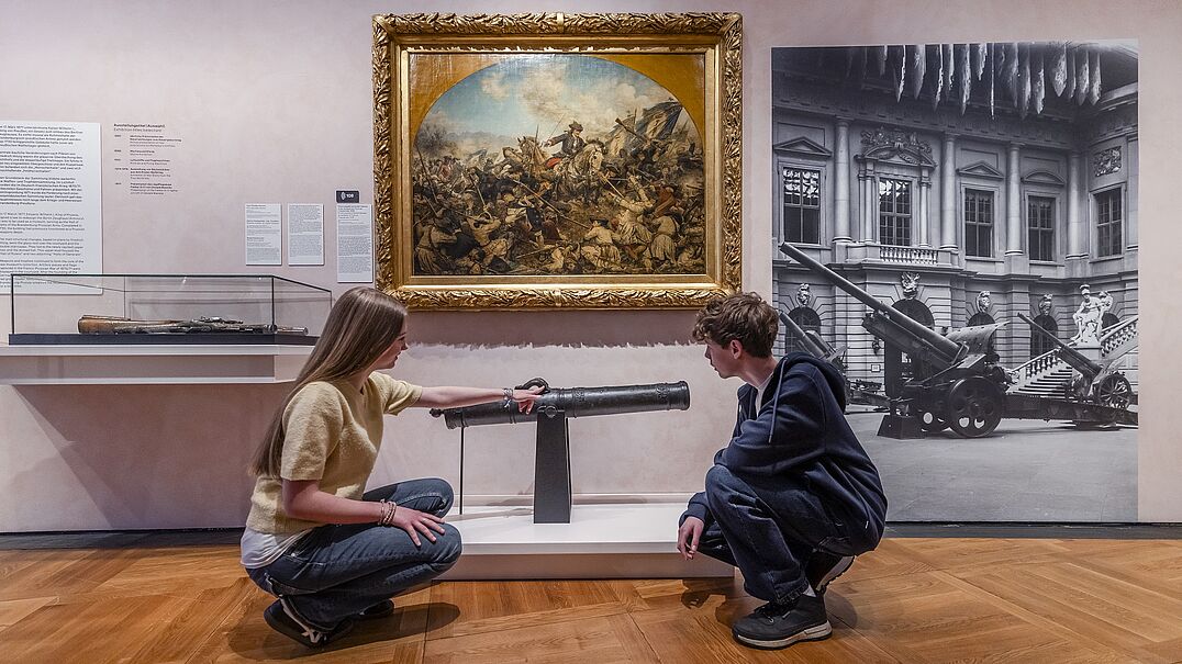 Zwei Kinder in einer Ausstellung und hocken vor einer Kanone. Sie schauen das Objekt interessiert an.