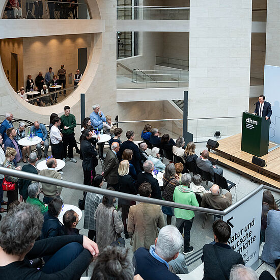 Viele Menschen stehen in einem Foyer. Eine Person spricht auf einer Bühne.