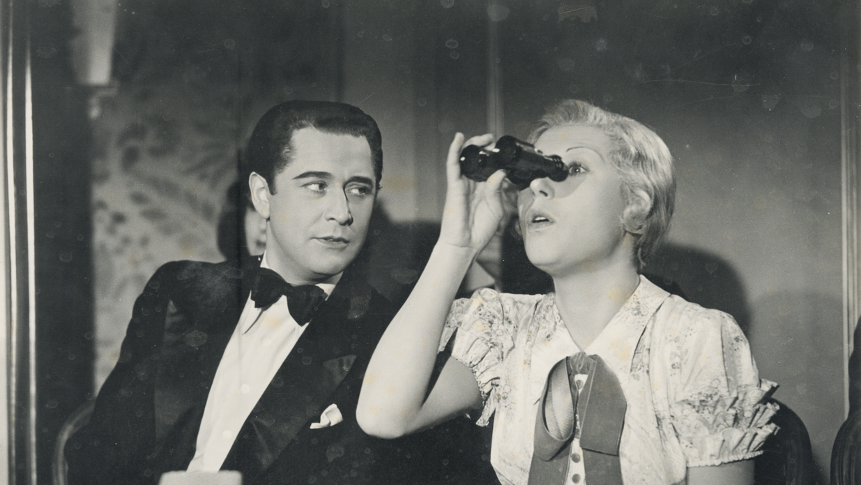 Eine Frau blickt in ein Fernglas und ein Mann sitzt neben ihr und betrachtet sie.