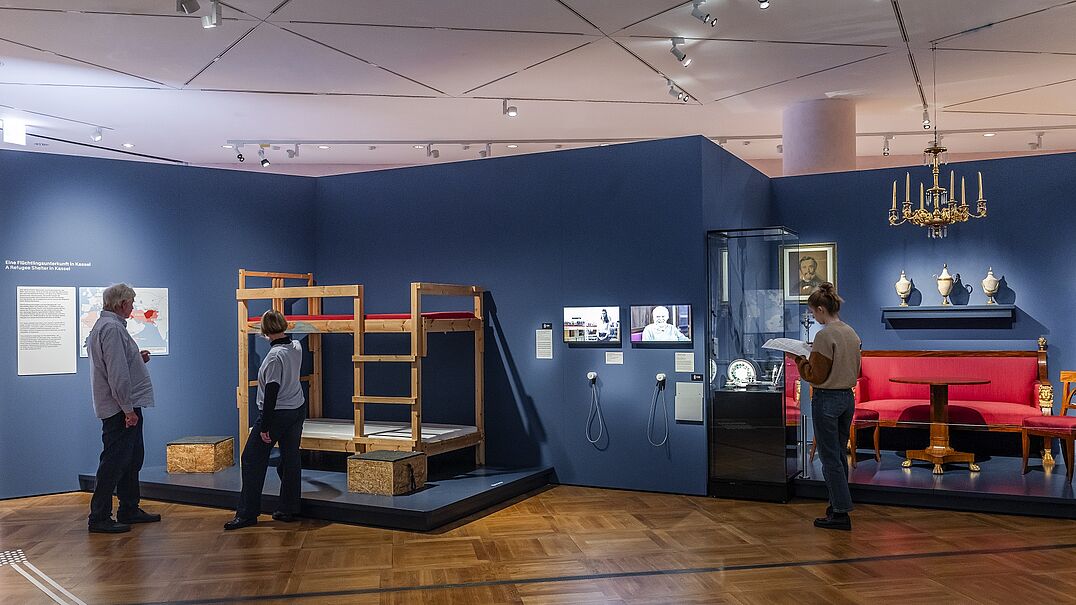 Menschen stehen in einer Ausstellung vor einem Bett aus einer Flüchtlingsunterkunft.